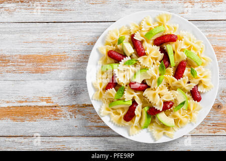 Leckere mini Salami bowtie Pasta warmer Salat mit Avocado Scheiben und Blätter Basilikum auf weiße Platte auf alten Holz- Hintergrund, Ansicht von oben Stockfoto