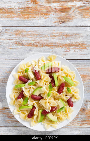 Leckere mini Salami bowtie Pasta warmer Salat mit Avocado Scheiben und Blätter Basilikum auf weiße Platte auf alten Holz- Hintergrund, Ansicht von oben Stockfoto