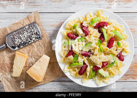 Würstchen Bow Tie Pasta warmer Salat mit Avocado Schichten und Basilikum bestreut mit Parmesan auf weiße Platte, mit Raspel und geriebenen Käse auf Holztisch, Stockfoto