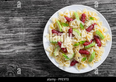Mini salami Bowtie Pasta warmer Salat mit Avocado Scheiben und Blätter Basilikum bestreut mit geriebenem Parmesan auf weiße Platte auf dunklen alten hölzernen Hinterg Stockfoto