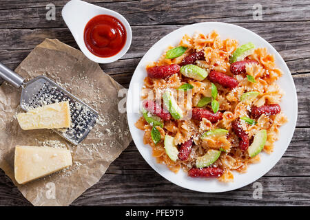 Würstchen bowtie Pasta warmer Salat mit Avocado Scheiben und Blätter Basilikum, mit Käse bestreuen auf weißem Schild auf Holztisch mit Reibe am Stück pap Stockfoto