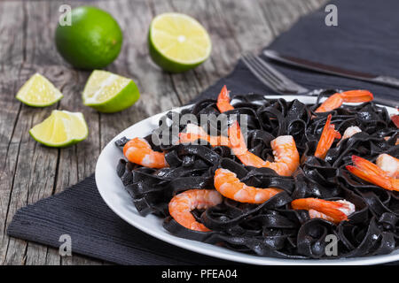 Lecker Tintenfisch schwarze Nudeln mit Garnelen auf einem weißen Teller auf graue Tabelle Matte mit Messer und Gabel, auf alten Holzplanken mit Scheiben von frischen Kalk auf Stockfoto