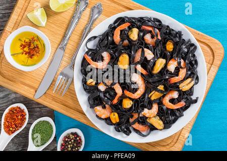 Lecker Tintenfisch schwarze Nudeln mit Riesengarnelen und Muscheln auf weißem Teller auf Schneidebrett mit Olivenöl und Gewürzen in Löffel auf alten hölzernen Hinterg Stockfoto