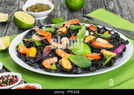 Köstliche schwarze Nudeln Salat mit Garnelen, Muscheln, frische grüne Spinat, Kopfsalat, Rucola auf weißem Teller am Tisch Matte auf dunklen Holztisch mit Kalk sl Stockfoto