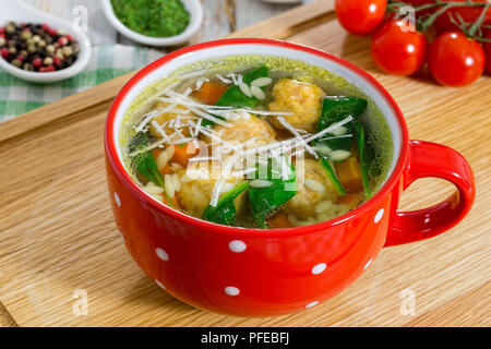 Köstliche italienische Hochzeit Hühnersuppe mit Frikadellen, kleine Nudeln risoni, Spinat und Gemüse in rot Schale auf Schneidebrett, Ansicht von oben, Schließen - Stockfoto