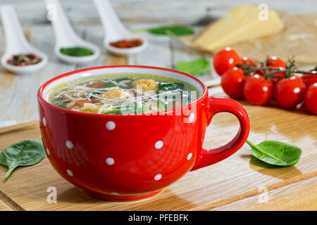 Köstliche italienische Hochzeit Hühnersuppe mit Frikadellen, kleine Nudeln risoni, Spinat und Gemüse in rot Schale auf Schneidebrett, close-up Stockfoto