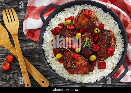 Reis mit gegrilltem Hähnchen Schenkel, rote Paprika, Grüne Oliven und Rosmarin in Gusseisen stewpan mit Küchentuch auf Holztisch, mediterrane Cu Stockfoto