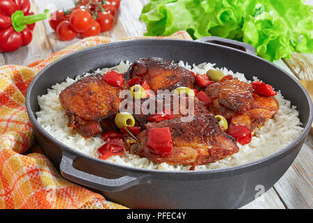 Reis mit gegrilltem Hähnchen Schenkel, rote Paprika, Grüne Oliven und Rosmarin in Gusseisen stewpan auf weißen alten Holztisch mit Küchentuch, Vegeta Stockfoto