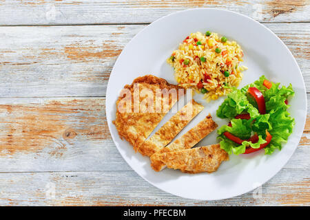 Knusprige Southern Fried Schweinekoteletts in Streifen geschnitten mit herrlich grünen Erbsen. Mais. Karotte Zwiebel würzigen Risotto und frischen grünen Salat auf weiße Platte Stockfoto