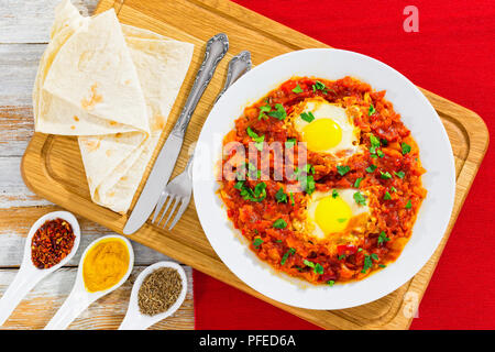 Shakshuka - Eier, Paprika, Chili, Tomaten und Gewürzen auf weiße Schale mit Fladenbrot auf Schneidebrett. Kreuzkümmel, Kurkuma, Pfeffer Flocken in Keramik Stockfoto