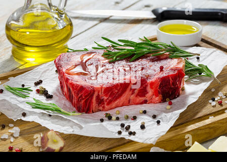 Rohes Rindfleisch Rib Eye frisches Fleisch Steak auf weißem Papier auf Schneidebrett mit Rosmarin gegossen mit Olivenöl, Knoblauch, koscheres Salz, Butter und Olivenöl Stockfoto