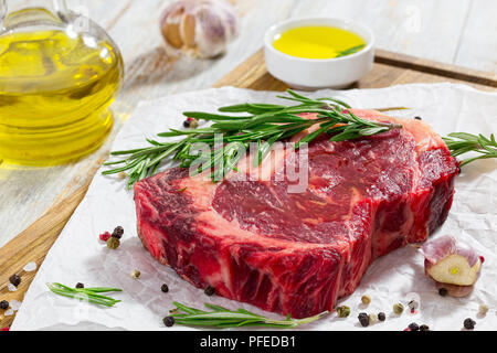 Rohes Rindfleisch Rib Eye frisches Fleisch Steak auf Holz Schneidebrett mit Rosmarin, Knoblauch, Nelken und Pfeffer mischen, ausgegossen mit Olivenöl zum Kochen vorbereiten, Stockfoto