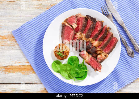 Saftiges Rindfleisch Steak medium mit Gewürzen in Scheiben schneiden auf weiße Platte mit gebratenem Knoblauch und Grünen frischen Spinat Salat. Messer und Gabel auf den Tisch Matte, v Stockfoto