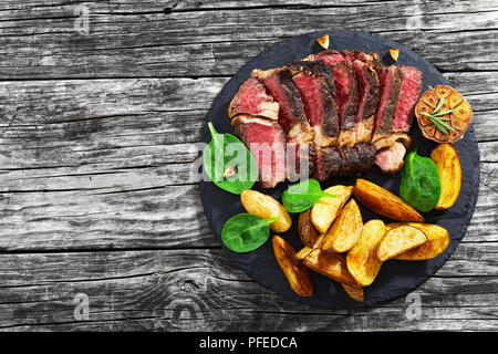 Rindfleisch saftiges Rib Eye Steak medium mit Gewürzen in Scheiben schneiden auf schiefer Platte mit gebratenem Knoblauch und Grünen frischen Spinat Salat. und gebratenen Kartoffelecken Stockfoto