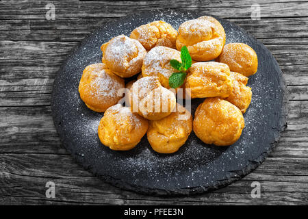 Leckere Windbeutel gefüllt mit Sahne auf schiefer Platte, Abstauben des pulverisierten Zucker, auf alten, dunklen Holztisch, Ansicht von oben, close-up Stockfoto