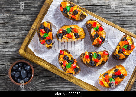 Crostini mit Stücken von Fisch gedünstet in Tomatensoße, Ringe von schwarzen Oliven, rote Paprika, auf weissem Papier auf Schneidebrett, Ansicht von oben Stockfoto
