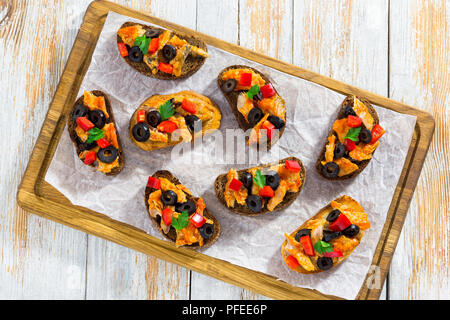 Bruschetta mit Stücken von Fisch gedünstet in Tomatensoße, Ringe von schwarzen Oliven, rote Paprika, auf weissem Papier auf Schneidebrett, Ansicht von oben Stockfoto