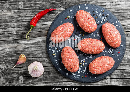 Rohes Fleisch Schnitzel auf schiefer Platte mit Meersalz und Mischung von Pfeffer auf alten, dunklen Holzbohlen mit getrockneten Chili und Knoblauch, Ansicht von oben Stockfoto