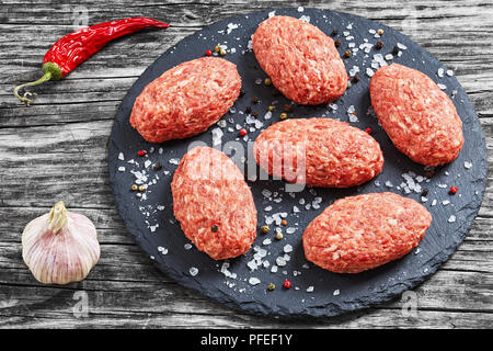 Frischem Boden rohes Fleisch Schnitzel auf schiefer Platte mit Meersalz und Mischung von Pfeffer auf alten, dunklen Holzbrettern, Ansicht von oben, close-up Stockfoto