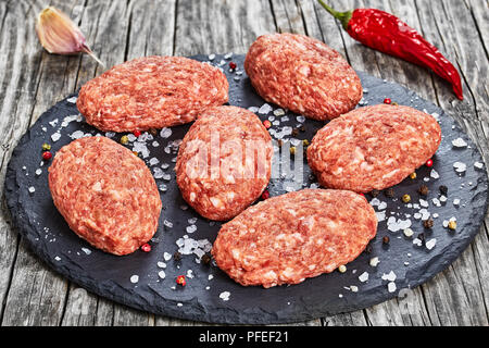Frischem Boden rohes Fleisch Schnitzel auf schiefer Platte mit Meersalz und Mischung von Pfeffer auf Altes, dunkles Holz mit getrockneten Chili und Knoblauch, Ansicht von oben, Stockfoto