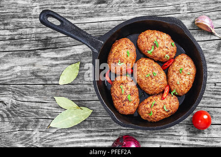 Saftige köstliche Fleisch Schnitzel in eisernen Pfanne bestreut mit Chili Stücke und grüne Kräuter. Tomaten, Lorbeerblätter und Zwiebeln auf alten rustikalen Tisch, Blick fr Stockfoto