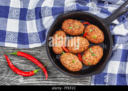 Saftige köstliche Fleisch Schnitzel in eisernen Pfanne bestreut mit Chili Stücke und grüne Kräuter. Tomate, Chili peppers auf alten rustikalen Planken, aus Abo anzeigen Stockfoto