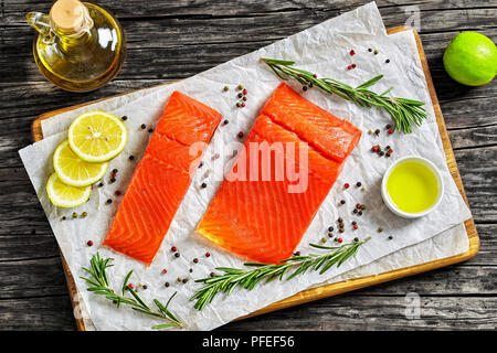 Lachsfilet mit frischem Rosmarin, Pfeffer ein Kalk Schichten auf Papier auf Schneidebrett. Flasche Olivenöl auf dunklen alten Holztisch, Ansicht von Abo Stockfoto