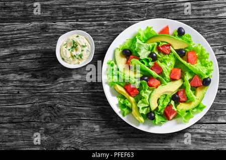 Lecker Salat von frischem Salat Blätter, Scheiben Avocado Brocken der gesalzenen Lachs und schwarze Oliven auf weiße Platte auf Holztisch mit hausgemachten Mayo Stockfoto