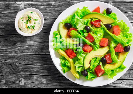 Lecker Salat von frischem Salat Blätter, Scheiben Avocado Brocken der gesalzene rote und schwarze Oliven auf weiße Platte auf Holztisch mit hausgemachten sa Stockfoto
