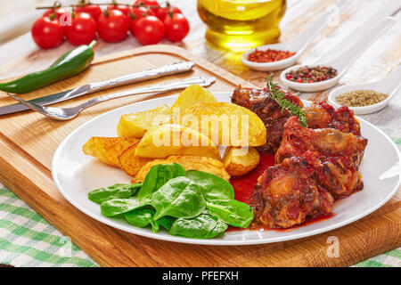 Ochsenschwanz in Rotwein geschmort mit Spinat Salat und gebratene Kartoffel auf weiße Platte auf Schneidebrett. Spice, eine Flasche Olivenöl und Tomaten Zweig auf Bac Stockfoto