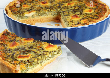 Quiche Florentiner mit Spinat, Käse, Eier und Gewürze in Schichten in Auflaufform, Stück Torte auf Papier auf weissem Holztisch mit Spachtel, Clos Stockfoto