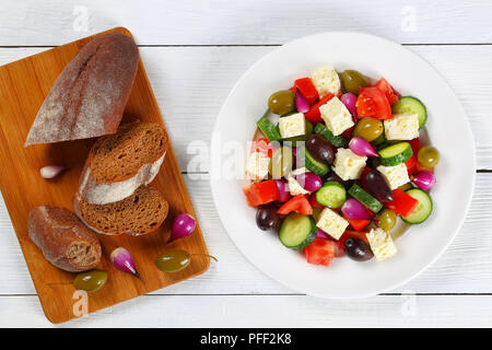 Leckere griechische Salat mit frischem Gemüse, Schafskäse grün und Kalamata Oliven, rote Zwiebel Zwiebeln auf weißen Teller am Tisch mit knusprigem Bauernbrot Stockfoto