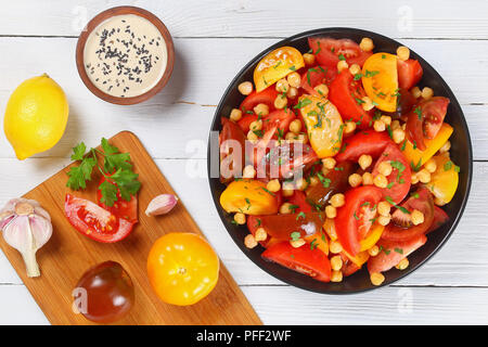 Kichererbse tomaten salat mit Petersilie in schwarz Schüssel, hausgemachte tahini Sosse bestreut mit schwarzem Sesam und eine Flasche Olivenöl auf weißer Tisch, in Stockfoto