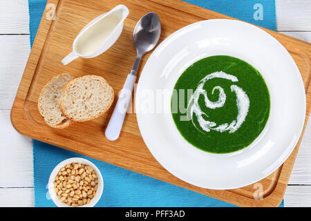 Kalten Sommer gesunde Suppe mit Spinat, grüne Zwiebeln, Brokkoli in weiten rim Suppenteller weiß Tisch, mit Sahne und Pinienkerne auf Schneidebrett, Ansicht von Stockfoto