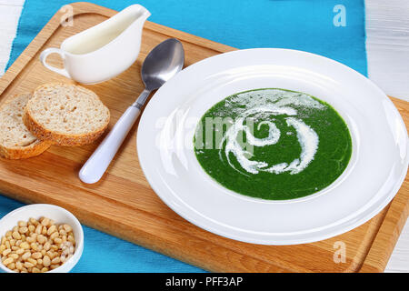 Sommer gesunde Suppe mit Spinat, grüne Zwiebeln, Brokkoli in weiten rim Suppenteller auf weißen Tisch mit Brot, Creme und Pinienkerne auf Schneidebrett, fr Stockfoto