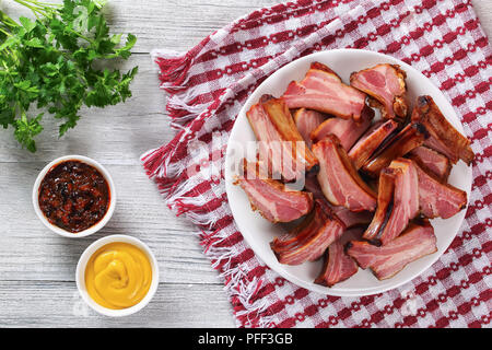 Lecker geräucherten Rippchen auf weißem Schild auf Holztisch mit Senf und Barbecue Sauce in Soße Boote, Ansicht von oben Stockfoto
