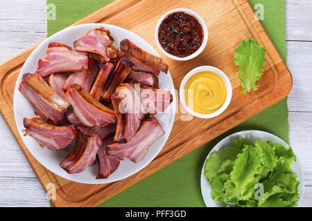 Lecker geräucherten Rippchen auf weiße Platte auf Holz Schneidebrett mit Senf und Barbecue Sauce in Soße Boote, frischem Salat Salat in der Schüssel auf Tabl Stockfoto