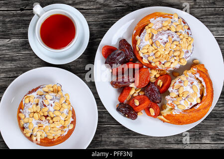 Hausgemachte köstliche frisch gebackenen Zimtschnecken gekrönt mit Erdnüssen auf Platte mit getrockneten Aprikosen, getrocknet Datum Obst/Tasse mit Tee auf Holz- Tabelle, Ansicht Stockfoto