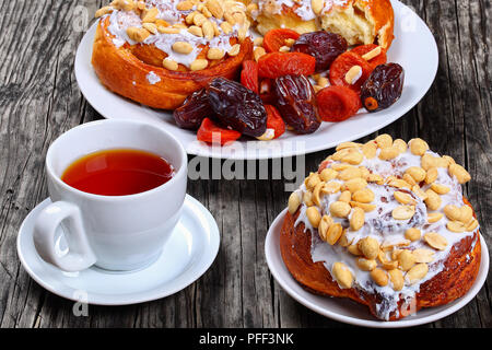 Hausgemachte köstliche frisch gebackenen Zimtschnecken gekrönt mit Erdnüssen auf Platte mit getrockneten Aprikosen, getrocknet Datum Obst/Tasse mit Tee auf Holz- Tabelle, Ansicht Stockfoto