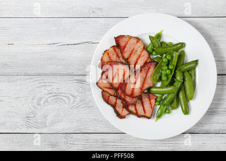 Köstliche gegrillte Fleischscheiben mit Streifen und gekochte Erbsen Pods auf weiße Platte auf hölzernen abblätternde Farbe Planken, Ansicht von oben Stockfoto