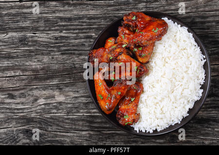 Reis mit köstlichen Honig Knoblauch Ingwer Glasierte Hähnchenflügel mit Petersilie in schwarz Schüssel auf dunklen Holztisch sprengte, Ansicht von oben Stockfoto