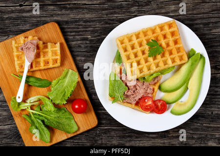 Leckere gesalzene belgische Waffeln Sandwich mit Avocado, Scheiben, Türkei Leber Pate, Salat, Spinat, Tomaten auf weiße Platte auf dunklen Holztisch, Ingr Stockfoto