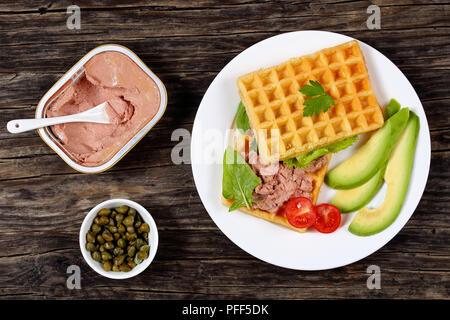 Leckere gesalzene belgische Waffeln Sandwich mit Avocado, Fleisch, Pilze und Leber Pate, Salat, Spinat, Tomaten auf weiße Platte auf dunklen Holztisch, Stockfoto