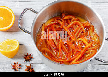 Hausgemachte kandierte Schalen Streifen von Orange und Zitrone in Sirup mit Zimt, Muskat und Anis Sterne im Topf auf alten hölzernen Tisch gekocht mit einer halben Orange Stockfoto