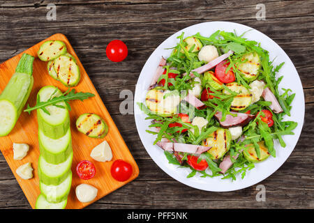 Köstliche gegrillte Zucchini, Mozzarella, Käse, Fleisch, Tomaten und Rucola Sommer Salat auf weiße Platte auf dunklen Tisch mit Zutaten auf ein Schneidbrett, Stockfoto