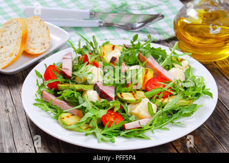 Köstliche gegrillte Zucchini, Mozzarella, Käse, Fleisch, Tomaten und Rucola Sommer Salat auf weiße Platte auf dunklen Tisch mit Brot und Olivenöl auf Backgr Stockfoto