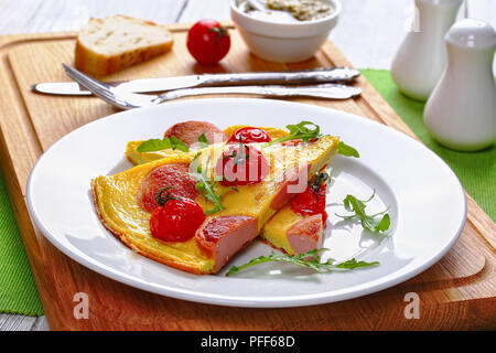 Teil der leckere Omelette mit Würstchen in Scheiben schneiden, Kirschtomaten und frische Rucola auf weißem Schild auf Schneidebrett mit Messer und Gabel, sa Stockfoto