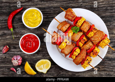 Leckere heiße gegrilltes Schweinefleisch mit Paprika und Teile von Maiskolben auf Bambus Spieße auf weiße Platte auf alten, dunklen Holztisch mit Senf und Tomaten Stockfoto