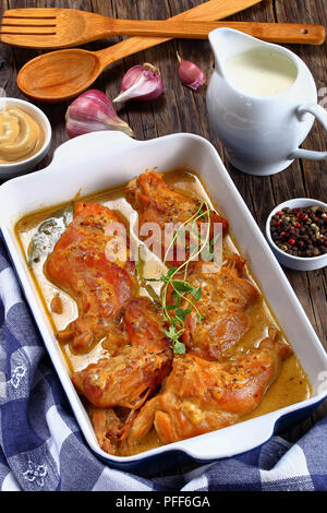 Appetitlich Kaninchen in Senfsauce gekocht oder lapin à la moutarde in der auflaufform auf Holztisch mit Creme Milchkännchen und Knoblauch auf Hintergrund, Klasse Stockfoto