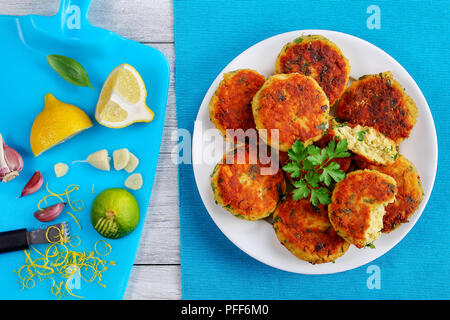 Köstliche frisch gebratenen Fisch, hausgemachte Kuchen auf weiße Platte mit Petersilie auf alten Holztisch, Keile von Limette und Zitrone auf ein Schneidbrett, klassisches Rezept, Stockfoto